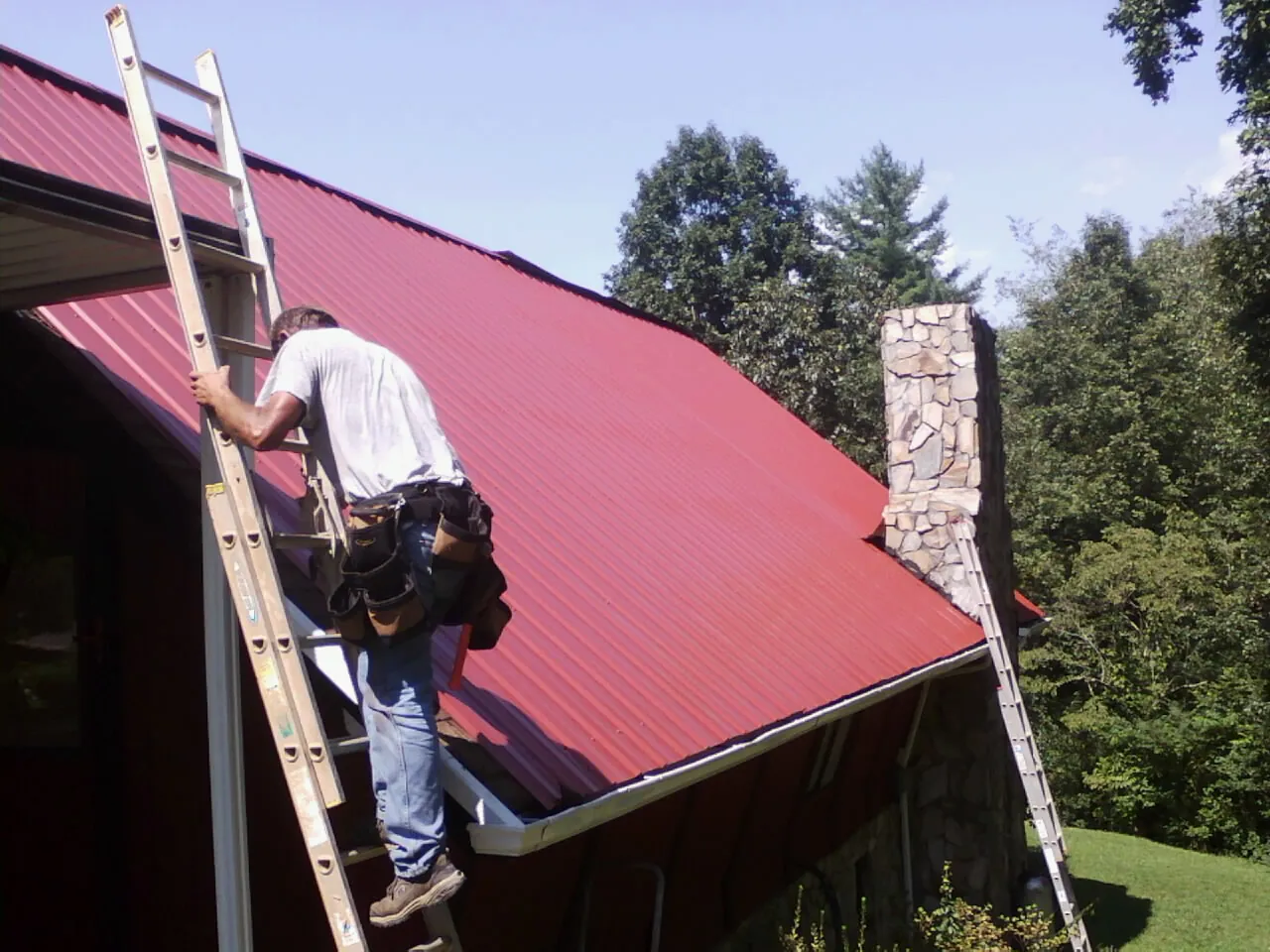 Roofer on a ladder beside a red metal roof and stone chimney — Soffit & Fascia Repair by Roofing Brothers in Brewster